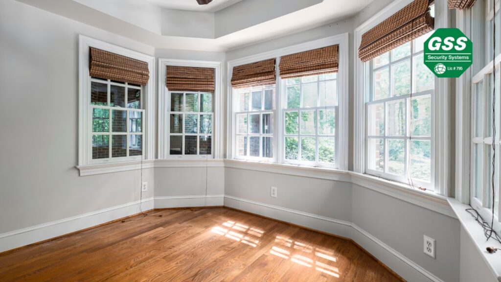 A bright and spacious bay window area in a home, featuring multiple large windows with natural light streaming in. The windows have woven blinds partially drawn, and the room has a hardwood floor with neutral-toned walls. A "GSS Security Systems" logo is visible in the upper right corner.