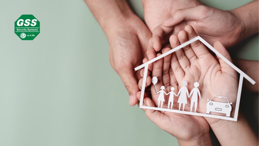 Overlapping hands cradle a paper cutout of a house with figures representing a family and a car, symbolizing the protection of home security systems.