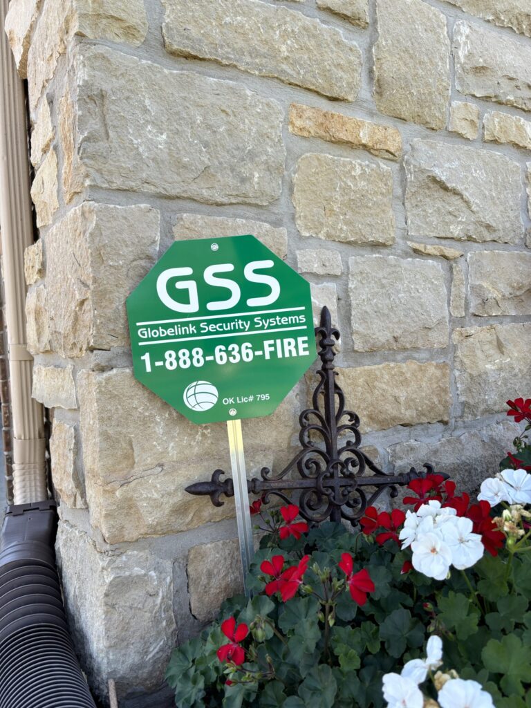 A garden sign for Globelink Security Systems with the logo "GSS" prominently displayed in green and white. Below the logo is the toll-free number "1-888-636-FIRE" and the Oklahoma license number "OK Lic# 795". The sign is planted in a garden with red and white flowers, against a backdrop of a stone wall and decorative ironwork.