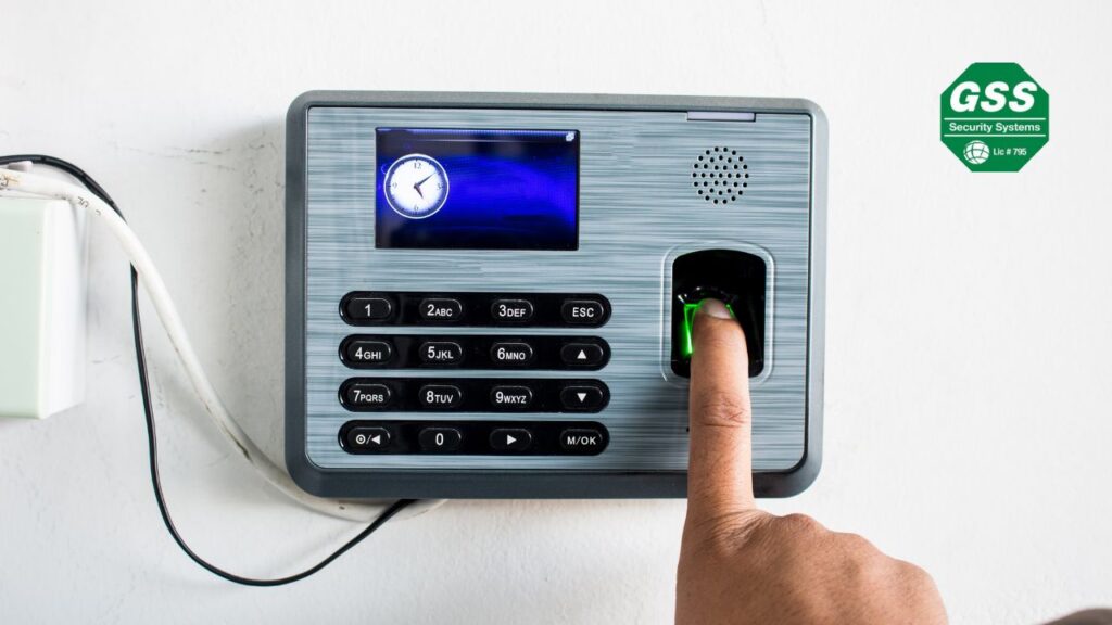 A person using a fingerprint scanner on an access control device mounted on a wall, with a digital clock display and numeric keypad. The GSS Security Systems logo is visible in the top right corner, emphasizing security and access management solutions for small businesses.